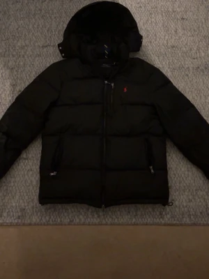 Svart pufferjacka från Polo Ralph Lauren - Svart pufferjacka från Polo Ralph Lauren med huva och röd broderad logga på bröstet. Jackan har två sidofickor med dragkedja och en extra bröstficka med dragkedja. Perfekt för kalla dagar och har en clean, stilren look.