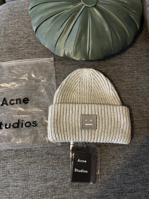 Grå stickad mössa Acne Studios - Snygg grå ribbstickad mössa från Acne Studios med klassisk smiley-patch framtill. Mössan är tillverkad i 100% ull och har en bred uppvikt kant för extra stilpoäng. Perfekt accessoar för att lyfta din look under kyliga dagar.