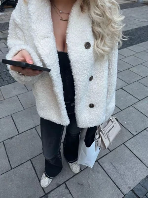 Vit teddyjacka med knappar - Säljer en fluffig vit teddyjacka med stora bruna knappar framtill. Jackan har en oversized passform och är långärmad, perfekt för dig som gillar mysiga och trendiga plagg. Materialet är mjukt och ger en cool look till din outfit.
