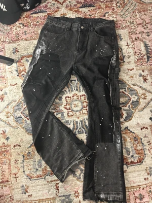 Svarta jeans med färgstänk och slitningar - Svarta jeans med coola vita färgstänk och slitna detaljer längs benen och vid bensluten. Byxorna har flera fickor, inklusive en sidoficka, och en rak passform. Materialet är klassisk denim och de har en avslappnad streetstil.