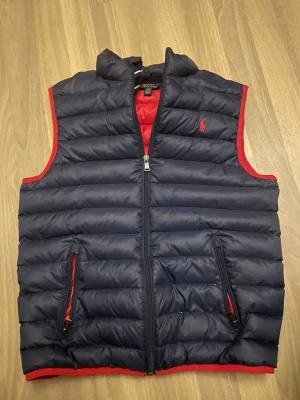Mörkblå dunväst från Polo Ralph Lauren - Snygg mörkblå dunväst från Polo Ralph Lauren med röd kant och röd logga på bröstet. Västen har quiltad design, två fickor med dragkedja och hög krage. Perfekt att slänga över en hoodie för extra värme. Stängs med dragkedja framtill.
