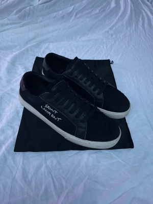 Svarta Saint Laurent sneakers - Svarta sneakers från Saint Laurent med vit logga på sidan. Skorna har snörning, rund tå och vit platt sula. Materialmix av skinn och textil ger en lyxig vibe. Perfekt för dig som gillar stilrena och exklusiva sneakers med streetkänsla.nästan helt nya och i super skick, pris är inte hugget i sten!