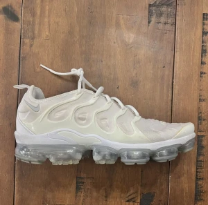 Nike Air Vapormax Plus  - Nike Air Vapormax Plus sneakers i helvitt med futuristisk design och genomskinlig sula. Ovandelen är i syntetmaterial med vågiga detaljer och snörning. Snygg och modern look med tydlig Nike-logga på hälen. Perfekt för dig som vill sticka ut med dina skor.