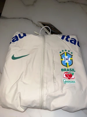 Vit Brasil Nike vindjacka med huva - Snygg vit vindjacka från Nike med Brasil CBF-logga och sponsormärken på bröstet och ärmarna. Jackan har huva, hel dragkedja och tryckta detaljer i blått, grönt och rött. Perfekt för dig som gillar fotboll och vill ha en sportig stil.