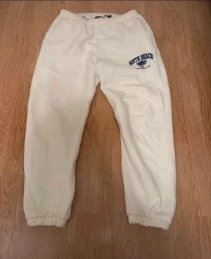 Vita mjukisbyxor från Lager 157 - Vita mjukisshorts från Lager 157 med blått tryck 'South Beach Padel Sports Club' på ena benet. Byxorna har resår i midjan och vid bensluten för en skön passform. Perfekta för chill eller sportiga dagar. Tyvärr har byxan en liten fläck framtill