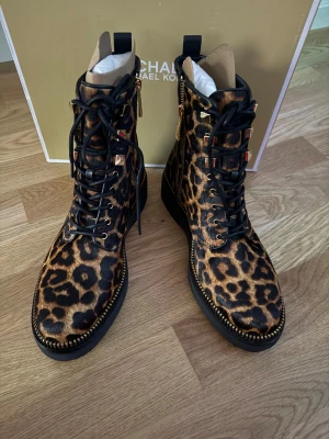 Leopardmönstrade boots från Michael Kors - Snygga boots från Michael Kors i leopardmönstrad mocka med svarta snören, guldfärgade detaljer och dragkedja på sidan. Grov platt sula och rund tå ger en cool look. Perfekta för dig som vill sticka ut med djurmönster och exklusiv känsla.