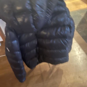 Svart Moncler-jacka 12 år - Säljer en svart Monclerjacka med glansig finish och markerade sömmar Maja. Klassisk look som funkar till det mesta och ger en cool vibe. Hål i luvan. 