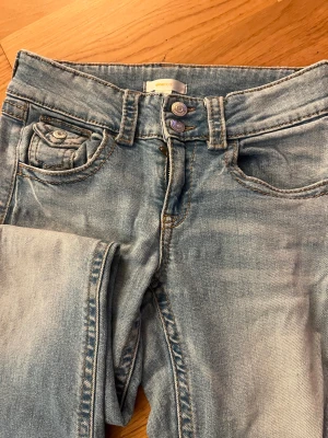 Bootcut jeans i ljusblå denim Gina Tricot - Snygga bootcut jeans från Gina Tricot i ljusblå tvättad denim. Klassisk femficksmodell med dubbla knappar i midjan och raka ben med lätt utsvängning. Perfekt för dig som gillar retrovibbar och bekväm passform.