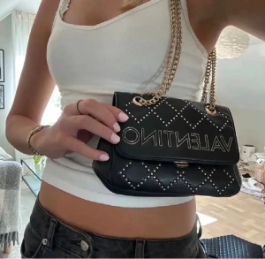 Svart axelväska från Valentino - Snygg svart axelväska från Valentino med quiltat mönster och silverfärgade nitar. Väskan har en kedjerem i guld och stor logga framtill. Aldrig använd då jag fick två liknande handväskor i 18 års present. Pris kan diskuteras!!