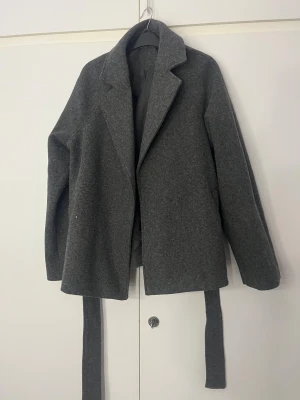 Grå omlott kappa med bälte - Snygg grå kappa med omlott-design och tillhörande bälte i midjan. Jackan har klassisk krage, raka långa ärmar och är tillverkad i ett ulligt material med slät insida. Perfekt för dig som gillar stilrena och tidlösa plagg. Andvänd ett få antal gånger, säljer den eftersom den är för stor. Fläcken går att få bort. Priset kan diskuteras. Helt ny kostar ca 500kr