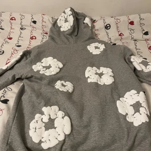 Denim tear hoodie grey  - Säljer en supermysig grå hoodie med vita blommor över hela tröjan. Perfekt för att hålla sig varm och stilren under kyliga dagar. Den har en klassisk passform med en stor ficka fram och en bekväm huva. Perfekt för både vardag och chill.
