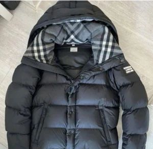 Svart pufferjacka från Burberry - Svart pufferjacka från Burberry med avtagbara ärmar, så du kan bära den som väst. Jackan har en huva med klassiskt rutigt Burberry-foder i grått och vitt, samt dragkedja och knappar framtill. Snygga detaljer med logotyp på ärmen och två fickor med dragkedja.