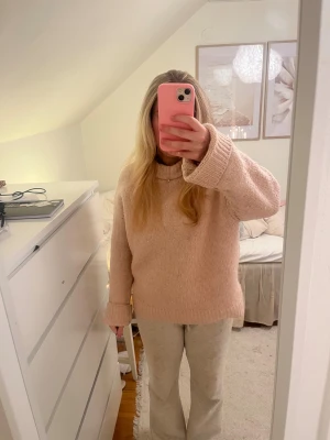 Beige stickad tröja från H&M - Mysig stickad tröja från H&M i en ljus rosa ton. Tröjan har en bred ribbad krage och långa ärmar med uppvikta muddar. Passformen är loose och materialet känns mjukt, perfekt för kyliga dagar. 