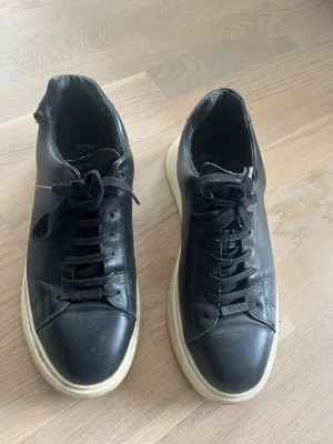 Svarta sneakers från BOSS i skinn - Boss Bulton. Ordinarie pris 2800kr. En liten skada på högra skon vid sidan av ankeln. Snygga svarta sneakers från BOSS med vit platt sula och rund tå. Skorna är tillverkade i skinn och har klassisk snörning med svarta bomullssnören. Diskret BOSS-logga på plösen och sidan av sulan. Perfekt för dig som gillar stilrena och tidlösa sneakers.