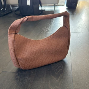 Brun flätad axelväska i skinnimitation - Säljer en snygg brun axelväska med flätad struktur och bred axelrem. Väskan har dragkedja upptill och ett extra fack med dragkedja på baksidan. Väskan är helt oanvänd med lappar och skyddsplast kvar. Säljer då den aldrig kommit till användning.