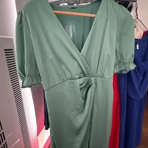 Grön omlottklänning med puffärm - Snygg ljusgrön omlottklänning med v-ringning och puffiga kortärmar. Klänningen har fina rynkdetaljer i midjan och en lätt glansig yta som ger en lyxig känsla. Perfekt för dig som vill sticka ut med en trendig och fräsch look.