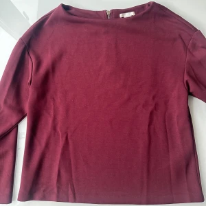 Vinröd långärmad topp med dragkedja - Vinröd långärmad topp i mjukt material med rund halsringning och snygg dragkedja baktill. Enkel och stilren design som passar perfekt till jeans eller kjol. Färgen är djup vinröd