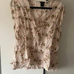 Blommig blus med snörning från Lindex - Supersöt blommig blus från Lindex i ljusrosa med gröna och röda blommor. Blusen har snörning framtill, v-ringning och är ärmlös. Perfekt för vår och sommar när du vill ha en luftig och romantisk look.