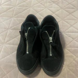 Svarta sneakers från Axel Arigato - Svarta sneakers i mocka från Axel Arigato med cool dragkedja framtill istället för klassisk snörning. Skorna har rund tå och platt sula, vilket ger en clean och modern look. Perfekta för dig som gillar minimalistisk stil med en twist.