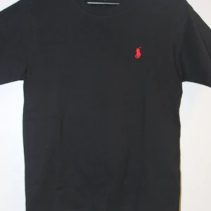 Svart t-shirt från Polo Ralph Lauren - Svart t-shirt från Polo Ralph Lauren med klassisk röd logobrodyr på bröstet. Modellen är enkel med rund hals och korta ärmar, perfekt för en clean och stilren look. Materialet är mjuk bomull som känns skönt mot huden. Helt nya aldrig använd. Pris kan diskuteras vid snabba affärer 