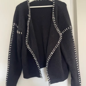 Svart kofta med vita detaljer - Svart stickad kofta med vita, grova sömmar längs kanterna och ärmarna. Koftan har v-ringning och två vita knappar framtill🖤🤍🖤🤍