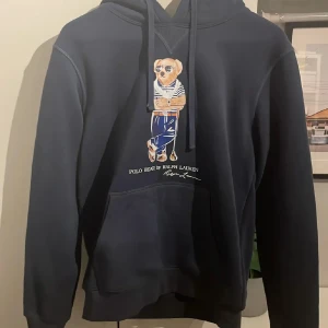 Mörkblå Polo Bear hoodie från Ralph Lauren - Säljer en mörkblå hoodie från Ralph Lauren med ikoniska Polo Bear-trycket på bröstet. Tröjan har huva med snörning, magficka och långa ärmar. Perfekt för dig som gillar klassisk streetstyle med en twist. Materialet känns mjukt och skönt mot huden. Perfket till alla kläder alltså passar den till allt. Hör av dig vid minsta fundering🤗.