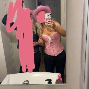 Rosa korsett med rosett och spets - Supersöt rosa korsett med glansig finish, dekorativ rosett framtill och vita spetsdetaljer längs kanterna. Knytdetalj vid bysten och knappar framtill. Perfekt för dig som vill sticka ut med en feminin och trendig look.