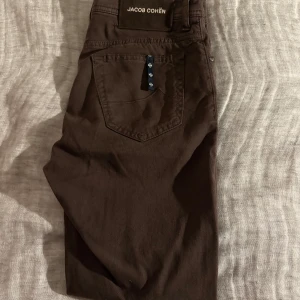 Jacob Cohën BARD, stl 31 - Säljer ett par bruna byxor från Jacob Cohën i storlek 31. Byxorna har klassisk femficksdesign, snygga detaljer med broderad logga och mönstrat innerfoder. Tillverkade i mjuk bomull med knappgylf och diskreta nitar vid fickorna. Har använts ca 5 ggr. De har sytts upp lite längst ner så att dem är lite kortare än nyköpta gjort på snyggt sett märks ej. Köptes för 4700 nya i Genève för 2 veckor sedan. Sitter straight legged och riktigt skönt material och relaxed passform.