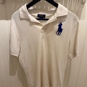 Vit polo t-shirt från Ralph Lauren - Vit polo t-shirt från Ralph Lauren med klassisk krage och korta ärmar. Stor blå broderad logga på bröstet ger en snygg kontrast. Tillverkad i mjuk bomull som är skön mot huden. Perfekt för dig som gillar stilrena och sportiga plagg.