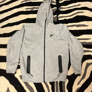 Grå Nike hoodie med dragkedja M - Snygg grå hoodie från Nike med svart logga på bröstet. Tröjan har hel dragkedja, två fickor med svarta detaljer och huva. Perfekt för chill eller träning, och har långa ärmar. Mjuk och bekväm i bomullsblandning.