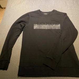 Svart sweatshirt från Hugo Boss - Svart sweatshirt från Hugo Boss i bomull med broderad logga framtill och en snygg matt detalj under texten. Tröjan har rund hals och långa ärmar, perfekt för dig som gillar stilrena och klassiska plagg med en modern touch.