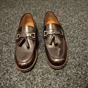 Vinröda loafers med tofsar och gulddetaljer - Snygga vinröda loafers i skinn med dekorativa tofsar och guldfärgade spännen framtill. Skorna har rund tå, platt sula och klassisk söm längs kanten. Perfekta för en dressad look med en twist. Insidan är ljusbrun och sulan är beige med diskret mönster. Aldrig använt.