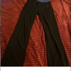 Svarta bootcut träningsbyxor BPC - Svarta träningsbyxor från BPC med blå midja. Byxorna har bootcut-modell och är tillverkade i ett stretchigt syntetmaterial som passar perfekt för träning. Snygg passform med låg midja och bekväm känsla. Perfekta för dig som gillar sportig stil.