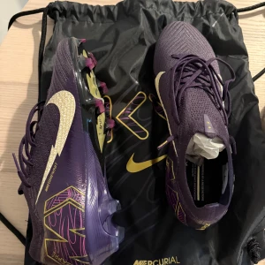 Nike Mercurial Mbappé fotbollsskor lila - Snygga Nike Mercurial fotbollsskor i samarbete med Kylian Mbappé. Skorna är lila med guldiga detaljer, vita swoosh-loggor och coola grafiska mönster. Ovandelen är i syntet och textil, med snörning och platt sula med dobbar för gräsplan. Helt nya och aldrig använda enbart testade. Storlek 42/5