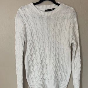 Vit kabelstickad tröja  - Vit kabelstickad tröja med klassiskt flätmönster och rund hals. Tröjan har långa ärmar och breda ribbade muddar vid ärmslut och nederkant. Perfekt för lager-på-lager och chill höst- eller vinterstil.