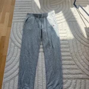 Grå mjukisbyxor från Champion - Grå mjukisbyxor från Champion med dragsko i midjan och sidofickor. Byxorna har en liten Champion-logga på vänster höft och är tillverkade i mjukt bomullsmaterial. Perfekta för chill eller träning.