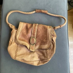 Brun axelväska i skinn med mönster - Säljer en brun axelväska i skinn med snyggt präglat mönster och dekorativt spänne i metall. Väskan har en bred axelrem som är lindad i skinn och detaljerade sömmar längs kanterna. Perfekt för dig som gillar vintagevibbar och unika accessoarer.