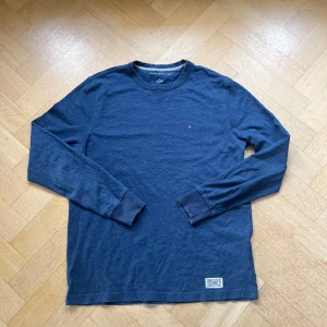 Blå långärmad tröja Tommy Hilfiger Storlek M - Mörkblå långärmad tröja från Tommy Hilfiger med klassisk rund halsringning och diskret broderad logga på bröstet. Tröjan är i mjuk bomull och har ribbade muddar vid ärmslut och hals. Snygg detalj med Hilfiger-märke nertill.