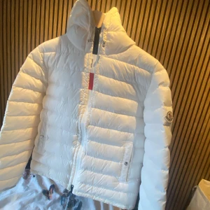 Vit Moncler rook down jacket  - Säljer en snygg vit dunjacka från Moncler med hög krage och huva. Jackan har klassisk pufferstil, dragkedja framtill och två fickor med knappar. Ikoniska Moncler-loggan sitter på ärmen. Perfekt för kalla dagar när du vill vara både varm och stilren. Den är i fint skick och den är äkta. Allt funkar på den såsom tag,nfc tag och sånt.  Storleken är 3 i Moncler men det är cirka som storlek M