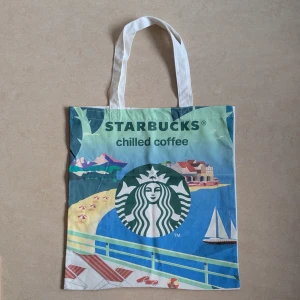 Starbucks tygkasse med strandmotiv - HELT OANVÄND TYGKASSE. Säljer en stor tygkasse från Starbucks med färgglatt strandmotiv på ena sidan och logga med text på andra. Väskan har långa handtag och är gjord i vit bomull. Perfekt för att bära böcker, laptop eller shopping. Snygg och unik accessoar för dig som älskar kaffe och sommarvibbar.
