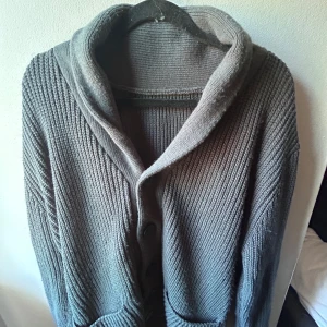 Grå stickad kofta med stora knappar från merley Stockholm  - Mysig grå stickad kofta med bred krage och stora knappar framtill. Koftan har två praktiska fickor och är ribbstickad för en cool struktur. Ett litet hål i en av fickorna.Perfekt för lager-på-lager och chill dagar. Passar dig som gillar en avslappnad och enkel stil. Sitter som L