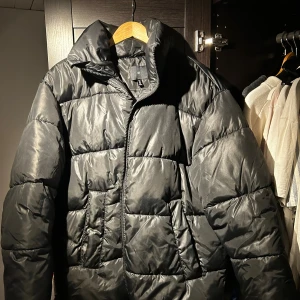 Svart pufferjacka med dragkedja - Snygg svart pufferjacka med hög krage. Jackan har dragkedja framtill och två stora fickor. Perfekt för kalla dagar. Enkel och clean design som passar till det mesta.