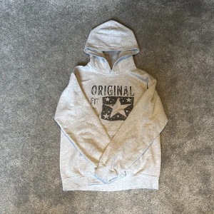 SCNDI - Ljusgrå SCNDI hoodie med svart tryck framtill där det står 'ORIGINAL' och en stor stjärna med mönster. Tröjan har huva, långa ärmar och ribbade muddar. Perfekt för en chill och avslappnad stil.                       Köpt på hemsidan för 899, mitt pris: 399.    OBS! Två små fläckar på mage som dock går bort i tvätt.