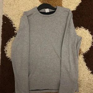 Grå långärmad tröja från NN07 - En stilren grå långärmad tröja från NN07. Tröjan har rund halsringning och är tillverkad i mjuk bomull, perfekt för lager-på-lager. Enkel och clean design som funkar till det mesta. Passformen är normal och materialet känns skönt mot huden.