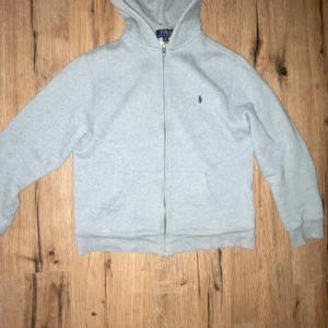 Grå hoodie från Polo Ralph Lauren - Snygg ljusgrå hoodie från Polo Ralph Lauren med dragkedja och klassisk blå logga på bröstet. Tröjan har huva, två fickor fram och är tillverkad i mjuk bomullsmix. Perfekt för chill dagar eller när du vill ha en clean och enkel look.