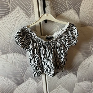 Zebramönstrad offshoulder topp - Snygg offshoulder topp från XOXO friends med svartvitt zebramönster. Modellen är croppad med puffiga korta ärmar och resår vid hals och midja för en cool och bekväm passform. Perfekt för dig som vill sticka ut med ett djurmönster!