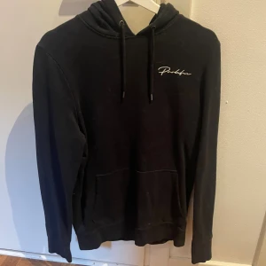 Svart hoodie från Peaklife - Svart hoodie från Peaklife med vit broderad logga på bröstet. Klassisk modell med huva, snörning och stor magficka. Perfekt för chill dagar och enkel att matcha med jeans eller joggers.
