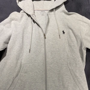 Grå hoodie från Polo Ralph Lauren - Ljusgrå hoodie från Polo Ralph Lauren med dragkedja, huva och snörning. Klassisk logga broderad på bröstet och två fickor framtill. Perfekt för en chill och stilren look.