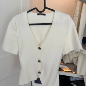 Vit v-ringad topp från Gina Tricot - Vit kortärmad topp från Gina Tricot med v-ringning och tre dekorativa guldiga knappar framtill. Toppens material känns mjukt och har en lätt stickad känsla, perfekt för en stilren och enkel look.