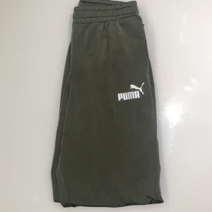Puma byxor - Snygga olivgröna byxor från Puma med elastisk midja och vid benslut. Byxorna har två sidofickor och en vit Puma-logga på låret. Perfekta för chill eller träning, tillverkade i mjukt och lätt polyestermaterial. 